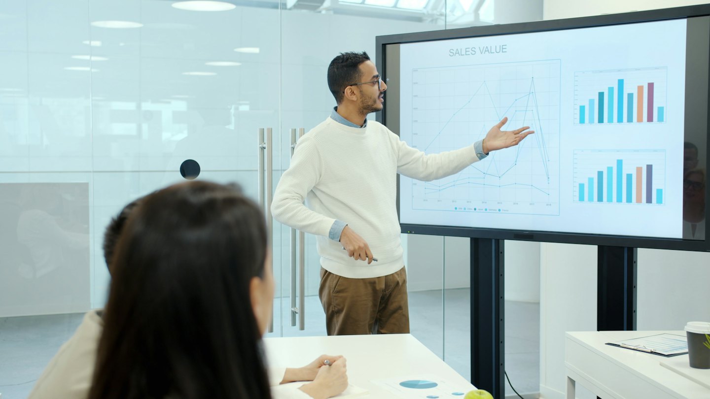 Hombre presentando datos de ventas en pantalla a colegas en una sala de reuniones.